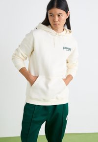 Sudadera con capucha color crema, con bolsillos frontales y un logo verde de "Converse All-Star"; combinada con pantalones verde oscuro. Textura suave.
