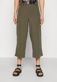 Culotte pants in olive green, featuring a relaxed fit, mid-calf length, smooth texture, and front pleats. Paired with black strapped sandals.