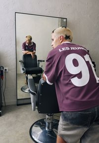 Black barber chair with ribbed upholstery, chrome base; person in a maroon jersey with "LES JEUNES" and "91," facing a mirror in a salon.