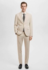 Bežna obleka iz gladkega blaga, ki vključuje enoredni suknjič z zareznimi klapi, v kombinaciji s tematskimi hlačami in črnimi čevlji.