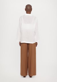Camicia oversize bianca in lino con maniche lunghe, abbinata a pantaloni larghi marroni. L'outfit presenta una vestibilità rilassata e una texture morbida.