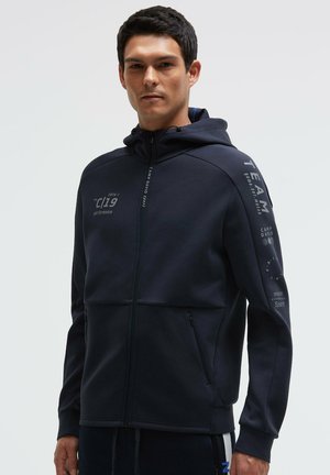 Man wearing a navy blue zip-up hoodie with hood, graphic text on chest and sleeve, standing against a plain light background.