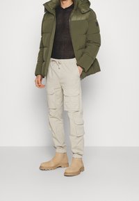 Olivově zelená prošívaná bunda, černý svetr s texturou, béžové kalhoty typu cargo s velkými kapsami a světle hnědé kotníkové boty s tlustými podrážkami.