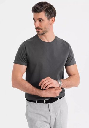 Graues kurzärmliges T-Shirt mit Rundhalsausschnitt, kombiniert mit hellgrauen Hosen und einem schwarzen Gürtel. Silberne Uhr am Handgelenk.