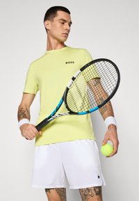 Neon gul kortermet treningstrøye med svart "BOSS"-logo, kombinert med hvite shorts. Holder en tenniskølle og en grønn tennisball.