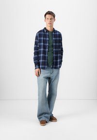 Plaid flannel shirt in navy and light blue over a green t-shirt, paired with light blue jeans and brown slip-on shoes.