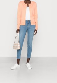 Ljus persikofärgad cardigan med rund halsringning och knäppning, i kombination med ljusblå jeans, vita sneakers och en mönstrad handväska.