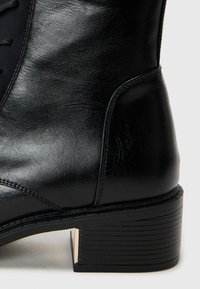 Black leather ankle boot with a smooth texture, round toe, lace-up design, and a low block heel featuring a metallic accent on the back.