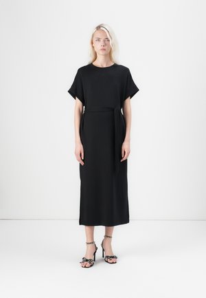 Black, ankle-length dress with short sleeves and a waist tie; smooth fabric; paired with black heeled sandals featuring polka dot bows.