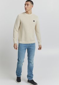 Suéter texturizado beige claro con cuello redondo, combinado con jeans desgastados azul claro y zapatillas negras, de pie contra un fondo neutro.