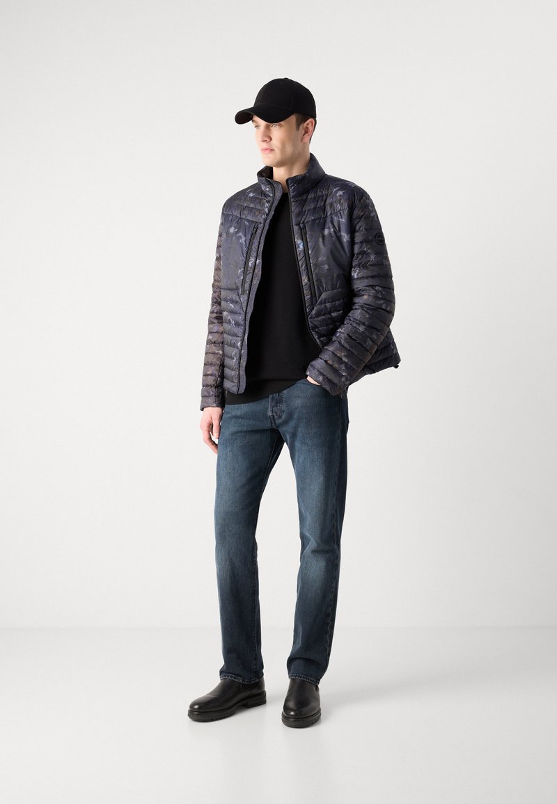 Puchowa, granatowa kurtka z kołnierzem stojącym i pikowanym wzorem, noszona na czarnej bluzie i niebieskich dżinsach, w zestawieniu z czarnymi butami i czapką.