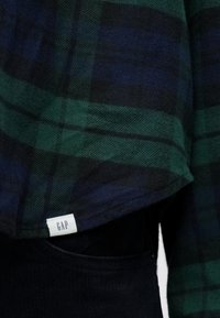 Πουκάμισο από φανέλα με πράσινο και μπλε καρώ, με στρογγυλεμένο τελείωμα και μικρή λευκή ετικέτα με το "GAP" κοντά στη ραφή στο κάτω μέρος.