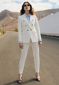 Biała dwurzędowa marynarka z złotymi guzikami, zestawiona z pasującymi spodniami i jasnoniebieską koszulą z kołnierzykiem. Noszona z czarnymi sandałami na obcasie.