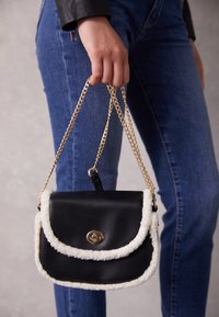 Svart läderhandväska med en krämfärgad pälsimitatkant, goldfärgad kedjeslinga och metalldetalj. Rektangulär form med invikbar klaffdesign.