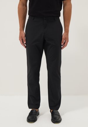 Mann trägt schwarze Slim-Fit-Hose und schwarze Ledermokassins, steht vor einem schlichten hellen Hintergrund.
