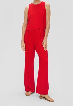 Femme portant une combinaison rouge sans manches à jambes larges et des sandales plates argentées, debout devant un fond clair uni.