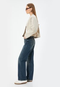 Beige cropped takki, jossa on leveät hihat, yhdistetty sinisiin leveälahkeisiin farkkuihin ja valkoisiin tennareihin. Asusteet sisältävät tummat aurinkolasit ja korvakorut.