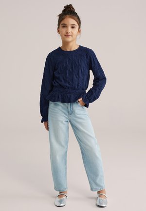 Tengerészkék, texturált, hosszú ujjú felső fodros szegéllyel, világoskék, bő szárú farmerrel és ezüst lapos cipővel párosítva.