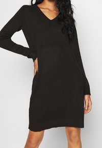 Robe pull noire en maille avec des manches longues, décolleté en V et poches latérales. Texture douce et coupe décontractée, arrivant au milieu de la cuisse.