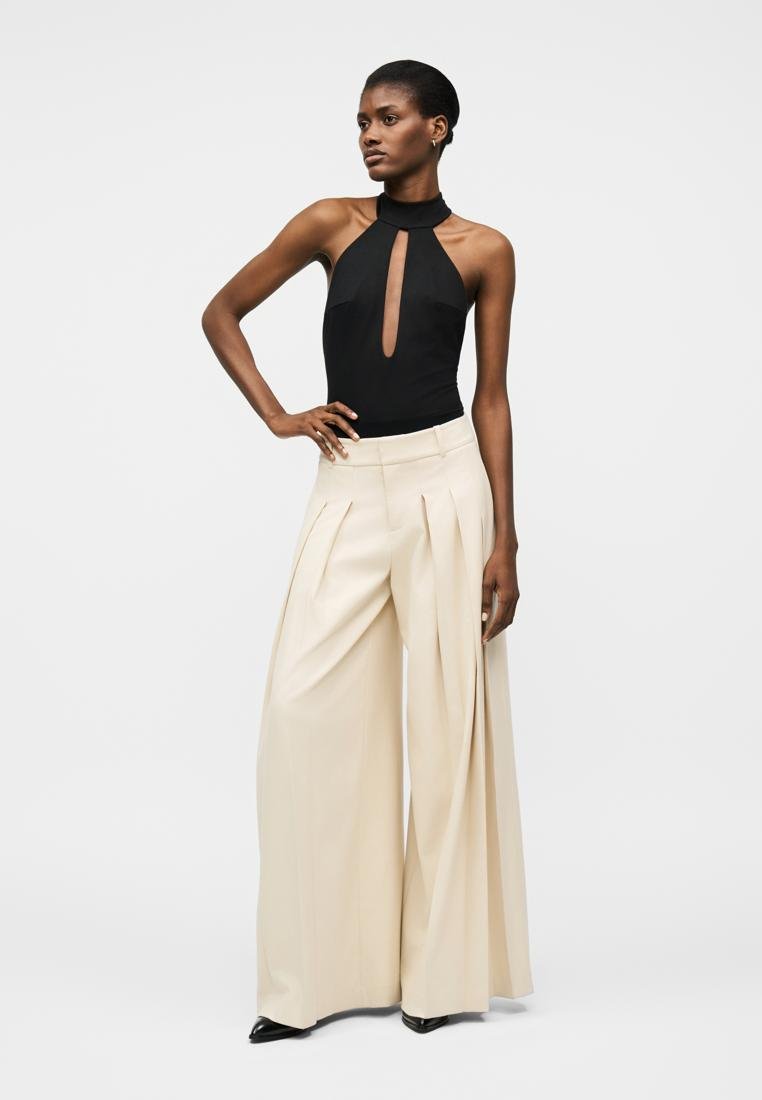 Femme portant un haut dos nu noir avec découpe frontale et un pantalon large crème, debout avec une main sur la hanche devant un fond uni.