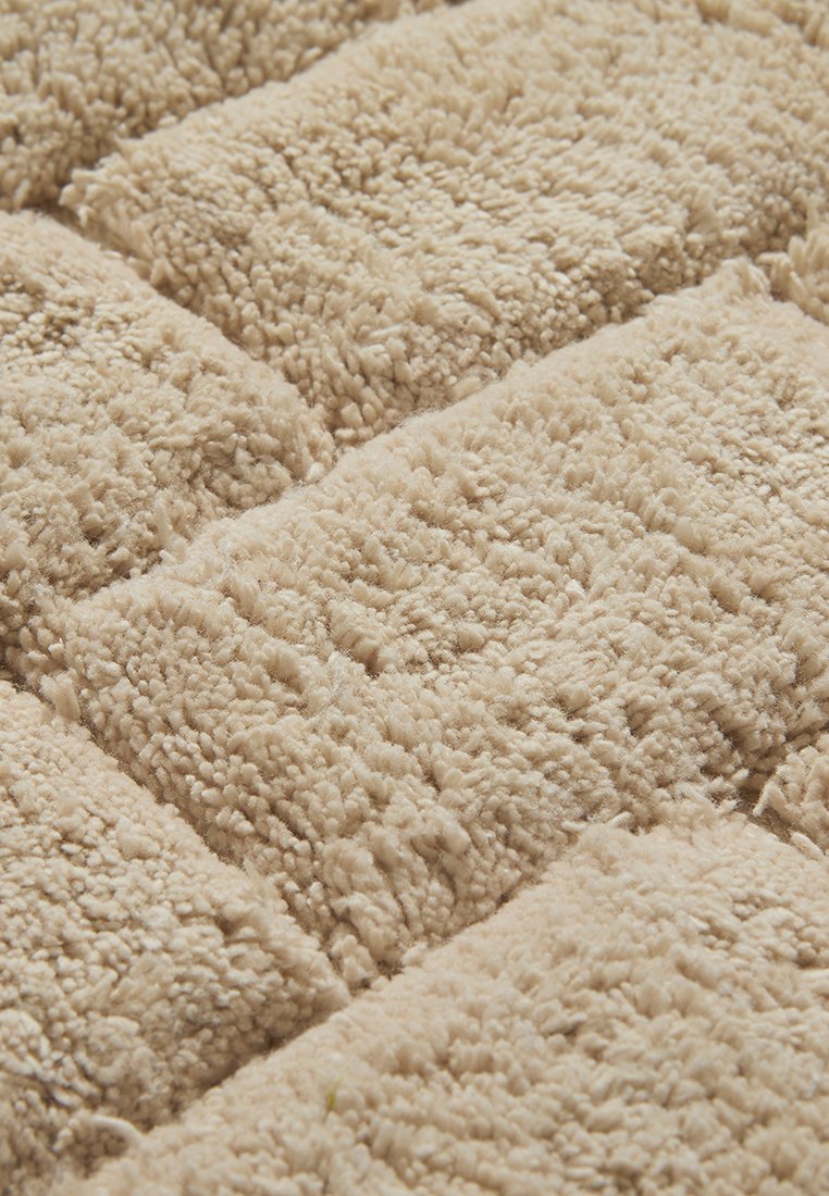 Beżowa tkanina pluszowa o teksturowanej, włochatej powierzchni ułożonej w pikowany wzór, prezentująca miękkie włókna i ciepły, zachęcający wygląd.