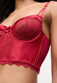 Lingerie in raso rosso con coppe in pizzo trasparente, spalline regolabili e bordino in pizzo a onde. Presenta un piccolo dettaglio a forma di fiocco al centro.