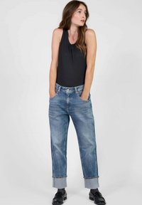 Bodysuit negro sin mangas combinado con jeans de tiro alto azul claro, con dobladillos enrollados y bolsillos laterales. La modelo está de pie con las manos en los bolsillos.