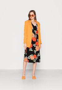 Vestido preto com padrões florais em laranja e azul, combinado com um blazer laranja. A modelo usa óculos de sol laranja e sapatos a condizer.