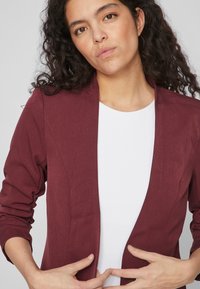 Bordowa marynarka o dopasowanym kroju, wydłużonych klapach i miękkim materiale; cechuje się podwiniętymi rękawami i minimalistycznym designem.