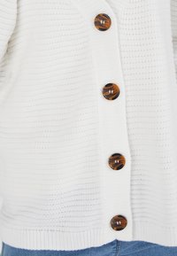 Cárdigan blanco de punto con tejido texturizado, seis grandes botones de mármol en tonos marrón y negro, y un diseño de corte relax.