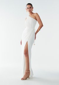 Vit, enfaldig klänning med en axel, draperad överdel och lårhög slits. Svalt, flytande tyg med knytning vid midjan. Guldiga remsandaletter syns.