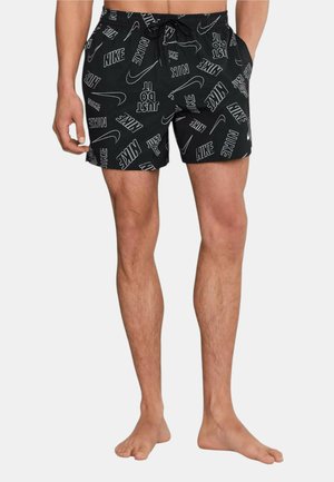 Uomo che indossa pantaloncini da bagno neri Nike con loghi bianchi e la scritta "Just Do It", in piedi scalzo davanti a uno sfondo semplice.