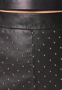 Gros plan sur un vêtement en cuir noir avec des clous argentés espacés régulièrement et une couture verticale visible au centre.