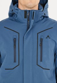Mann trägt eine blaue Jacke mit Kapuze, zwei vertikalen Reißverschlusstaschen auf der Brust und schwarzen Zierdetails auf der Vorderseite.