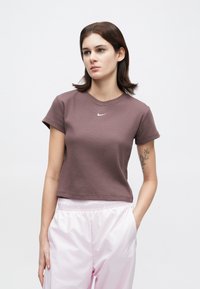 Νεαρή γυναίκα με κοντά καστανά μαλλιά, φορώντας ένα μωβ κοντομάνικο μπλουζάκι Nike και ανοιχτό ροζ αθλητικά παντελόνια, στέκεται με το ένα χέρι στην τσέπη.