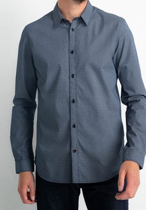 Homme portant une chemise à manches longues boutonnée avec un petit motif géométrique dans des tons bleus et un pantalon noir.