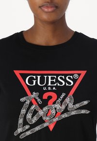 Crna majica s crvenim trokutastim logotipom. Bijeli natpis "GUESS" s ukrasima od kristala u obliku "Ikone" u sredini. Okrugli izrez.