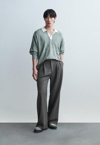 Νεαρός άνδρας στέκεται με τα χέρια στις τσέπες, φοράει ανοιχτοπράσινο πλεκτό πόλο με υφή, φαρδιά γκρι παντελόνια, λευκά αθλητικά παπούτσια και κοντό κολιέ.