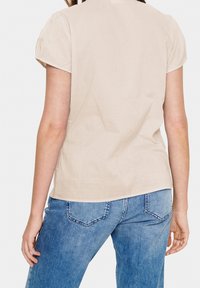 Helles beige Kurzarmoberteil mit lockerer Passform, ausgestattet mit einem runden Halsausschnitt und glatter Textur, kombiniert mit blauen Jeans.