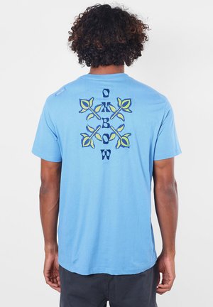 Homme aux cheveux bouclés portant un t-shirt bleu clair avec un motif floral jaune et bleu et le mot "Oxbow" dans le dos.
