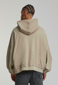 Sweat à capuche beige avec une coupe ample, présentant un ourlet texturé et des accents côtelés. Le tissu semble doux et légèrement brossé.