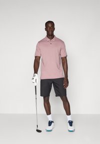 Chemise polo rose clair à manches courtes avec trois boutons, associée à un short noir, des chaussettes blanches et des chaussures de golf à accents bleus. Un club de golf tenu dans une main.