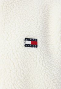 Hvid fleece-stof med en struktureret overflade. Har et sort mærke med "TOMMY" over et rødt og hvidt rektangulært logo og "JEANS" nedenfor.