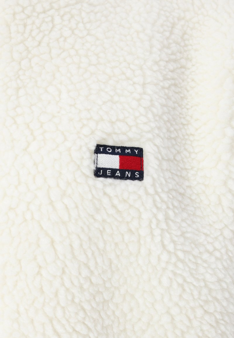 Tissu en polaire blanc avec une surface texturée. Présente une étiquette noire avec "TOMMY" au-dessus d'un logo rectangle rouge et blanc et "JEANS" en dessous.
