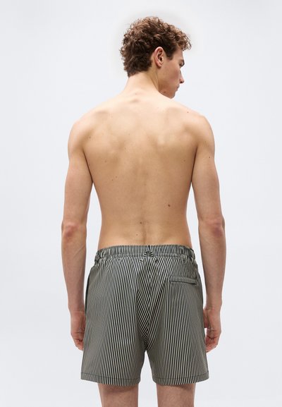 Homme vu de dos portant un short de bain à rayures verticales noires et blanches sur un fond clair uni.