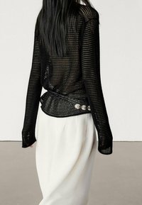 Woman with long dark hair wearing a black mesh long-sleeve top with silver buttons and a white skirt, shown from the back.