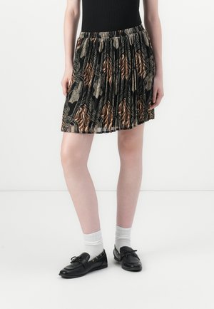 Jupe plissée noire avec un motif de feuilles colorées dans des nuances de marron et beige. Ceinture élastique. Portée avec des chaussures noires et des chaussettes blanches.
