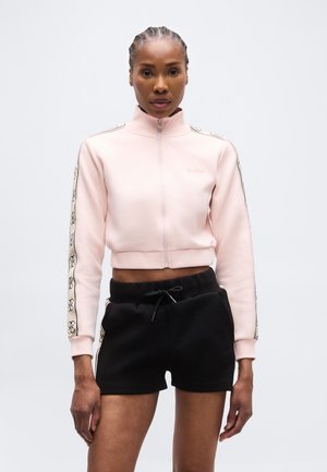 Femme de face portant une veste courte zippée rose clair avec des rayures à motifs sur les manches et un short noir à cordon de serrage.