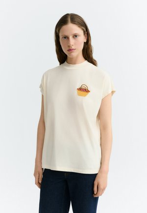 Femme à la peau claire et aux cheveux bruns portant un haut crème à manches courtes avec un petit panier de pique-nique brodé sur la poitrine et un pantalon foncé.