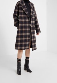 Abrigo largo de cuadros en colores oscuros con acentos en beige y rojo; presenta un cuello con muesca y un diseño cruzado. Llevado sobre una falda a cuadros.